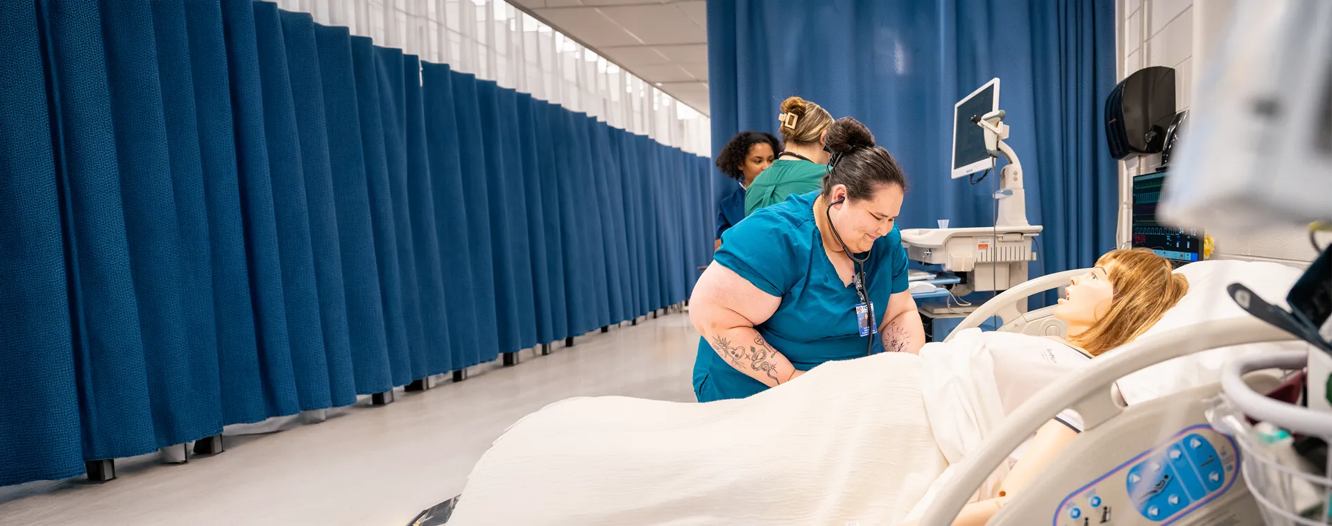 Student in scrubs providing care to a patient mannequin in a hospital training room with privacy curtains.