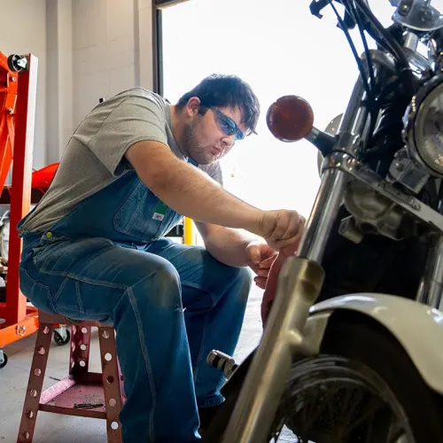 Student seated beside a motorcycle, using a hand tool during maintenance work in a training lab.
