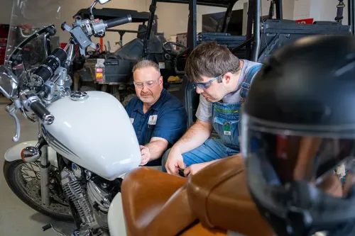 Instructor and student examining a motorcycle in a powersports lab with utility vehicles in the background.