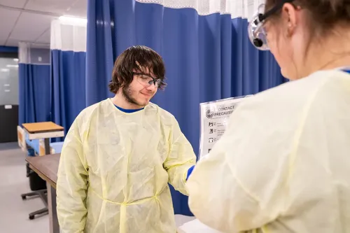 Students wearing protective gowns and goggles reviewing a contact precautions sign in a clinical lab.